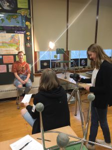 a high school student uses a video camera while another conducts an interview with a third student in a high school science classroom