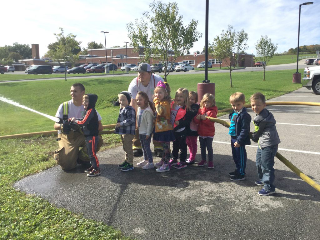 a firefighter helps a pre-k student spray a fire hose as the classmates hold up the hose behind them outside of an elementary school