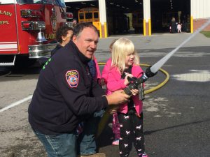 firefighter helps a pre-k student spray a fire hose outside of a school transportation building