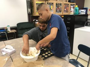 two students place batter from a bowl into a muffin pan at a table in a school classroom