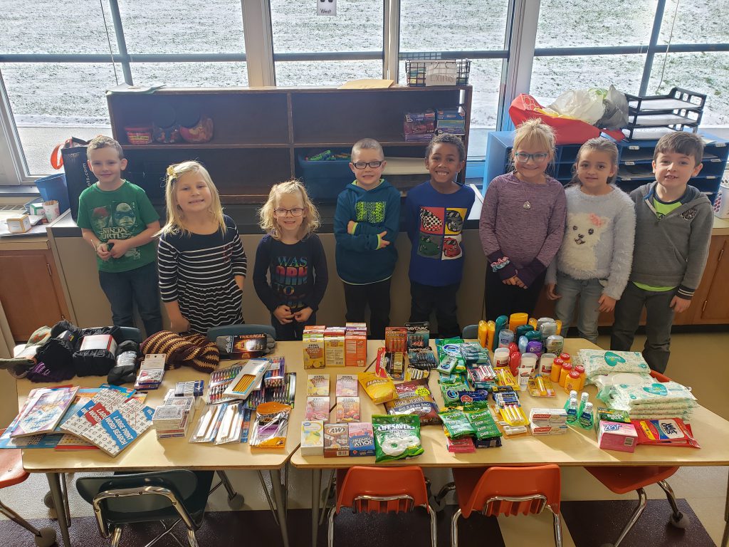 eight first grade students stand behind a table filled with donated items such as pens, candy, puzzle books and personal care items