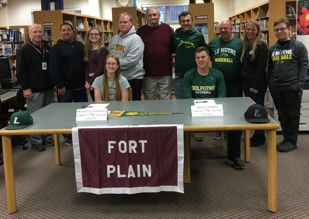 Two high school students seated at a table with a "Fort Plain" banner before a group of families, friends and educators 