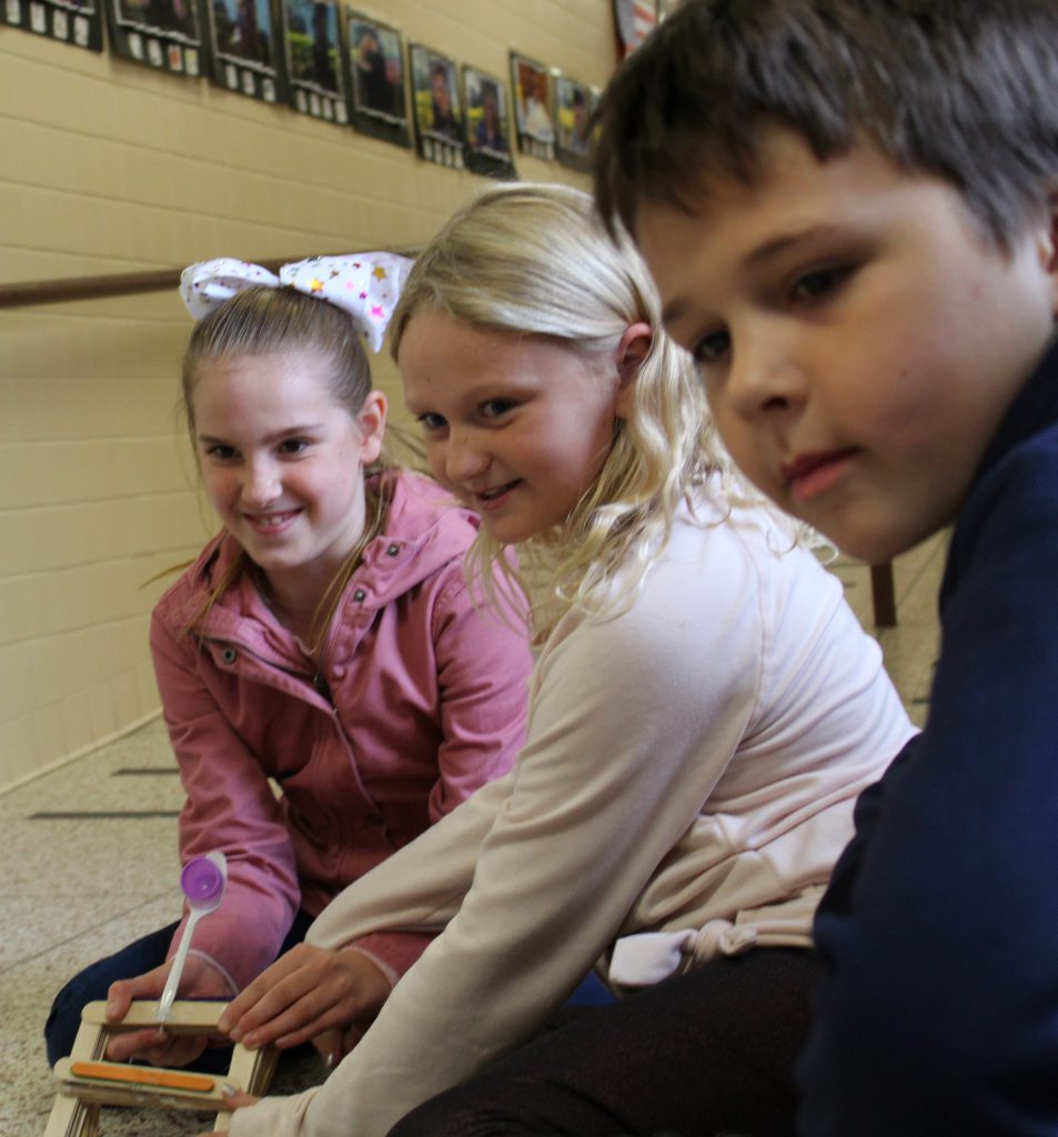 three students seated on the floor of a school hallway watch as a catapult launches a marshmallow pumpkin