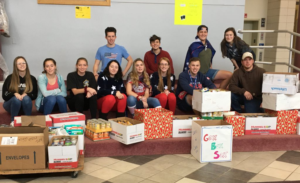 group of high school students sit in a school foyer with boxes of canned goods