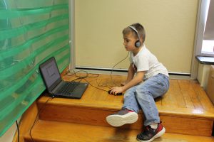 student sits on the top of a stairway landing while wearing headphones and clicking a computer mouse while using a laptop