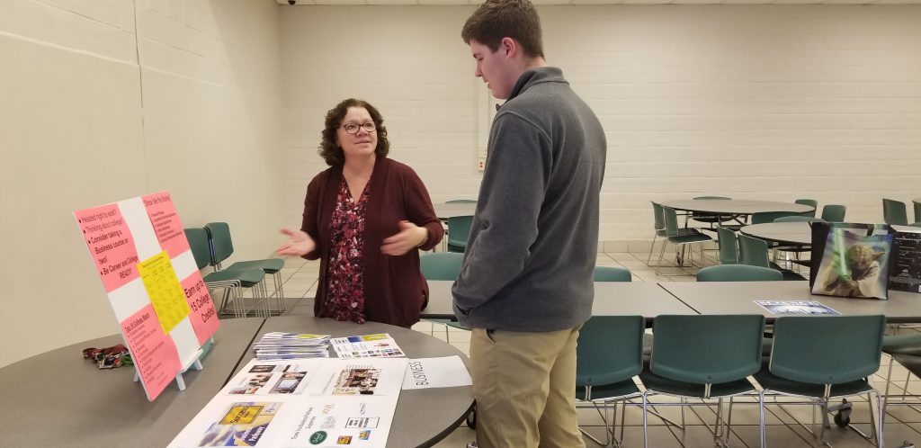 teacher talks to a high school student at a table in a cafeteria