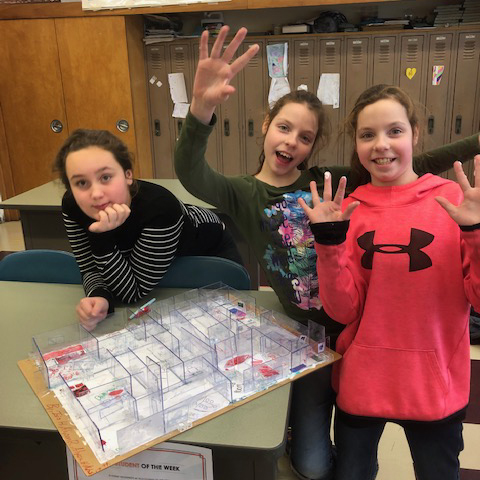 three elementary students stand next to a model home 