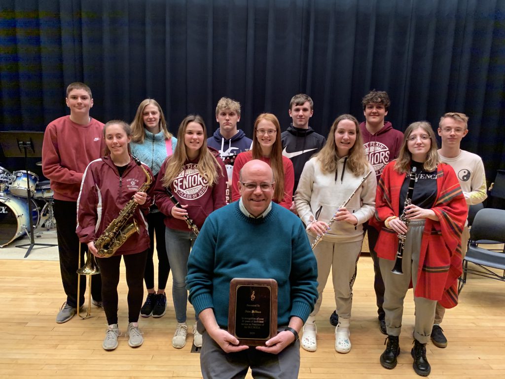 band director is seated on stage in school auditorium while student musicians hold instruments behind him. 