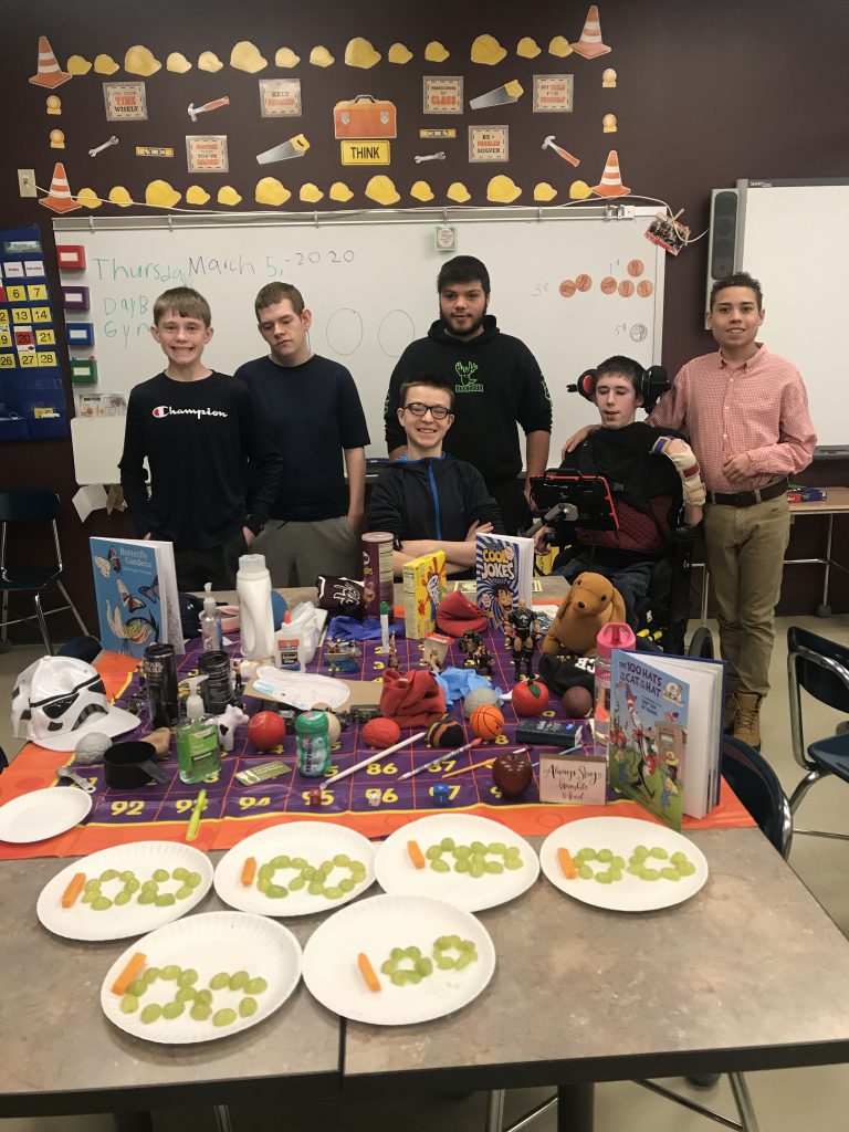 group of students stand before a table topped with plates of grapes and cheese sticks formed in the number 100