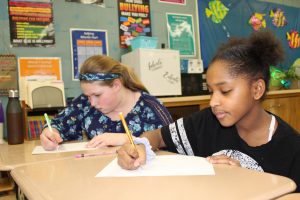 two students seated at desks use pencils to write on paper