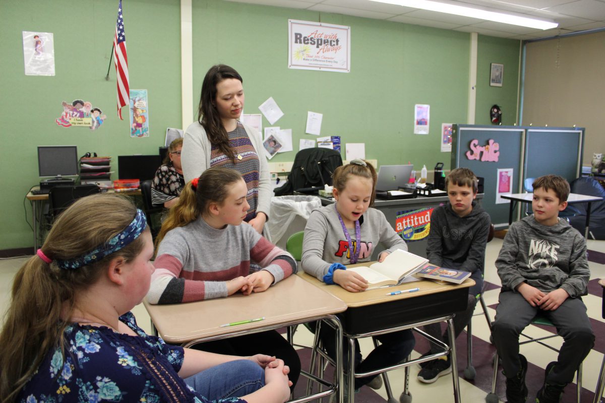 student reads from a book while sitting in a circle of students in a school classroom