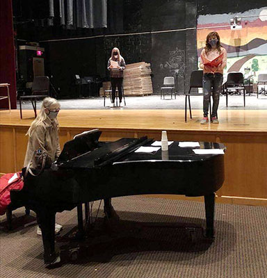 students reading scripts on stage while teacher plays the piano