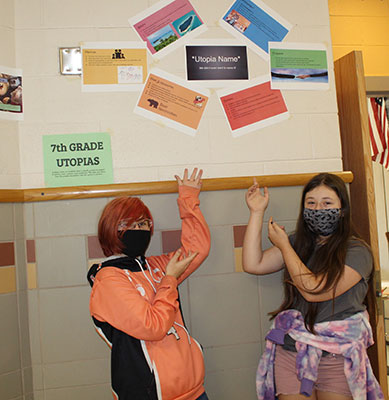 2 students point to their Utopia Projects posted on the wall above them