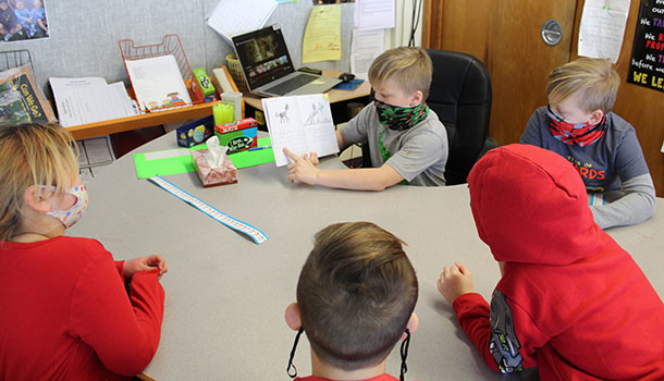 student holding book and reading to other students at a table