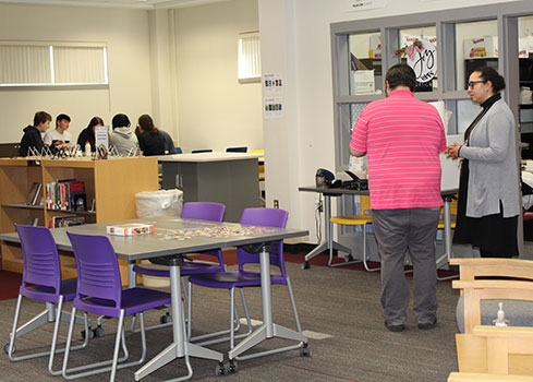 distant view of moveable tables, with students seated around one, and 2 adults talking in the foreground