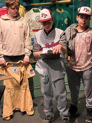 student holding clipboard, dressed in baseball gear while other students look on