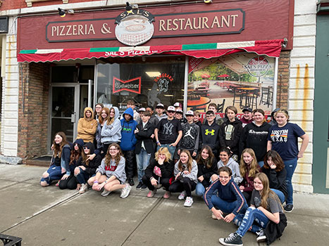 group of 7th grade students standing and sitting outside a pizzeria