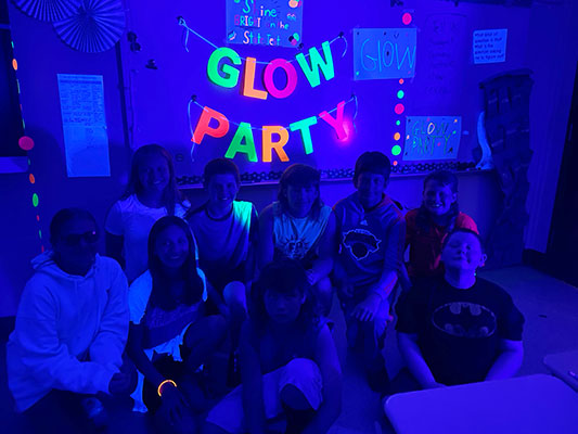students in a group in a blue-light-lit dark room with Glow Party sign