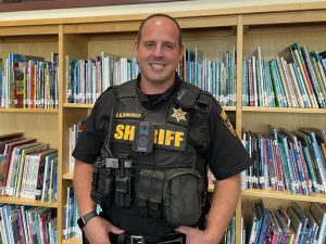 Deputy Sheriff Nicolas Manginelli, who has close cut brown hair, is wearing a black sheriff's uniform with the word "sheriff" in yellow across the chest, and gold sheriff's badge, stands in front of book cases full of picture books and looks at and smiles for the camera. 