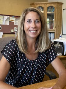 Fort Plain Central School District Superintendent Lauren Crisman, who has long blond hair and is wearing a printed short sleeved black blouse, sits at a wooden table in an office space and is looking at and smiling for the camera.