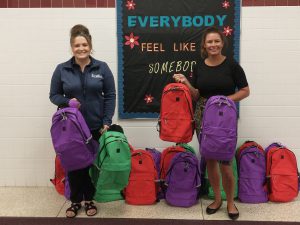 Harry Hoag Elementary school teachers, who are looking at and smiling for the camera, hold and stand in front of a row of colorful backpacks. 