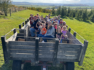 A group of Fort Plain first graders and teachers sit together in a wooden wagon at Bellinger's apple orchard. They are gathered close together and all look at and smile for the camera. It is a clear, sunny fall day and green grass, trees and mountains appear in the background. 