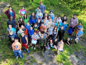 A group photograph taken from above of the fifth grade team at Harry Hoag Elementary. Students and teachers are all wearing hiking clothing and standing on bright green grass with rocky patches. 