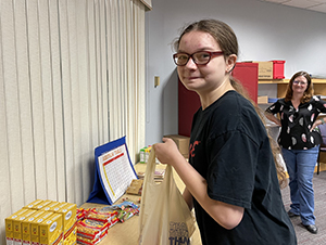 A high school student, who has long brown hair pulled back into a ponytail and is wearing eyeglasses and a short sleeved black t-shirt, works to fill a shopping bag with food items from a table. The student has turned and looks at and is smiling for the camera.