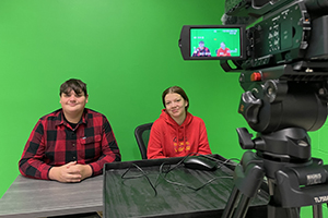 High school students Jacob Herringshaw and Madison Stuart sit at a table in front of a wall that is painted bright green. Both are looking at and smiling for the camera. In the foreground is a video camera. Jacob and Madison can be seen through the camera's preview screen. 