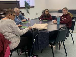 Two elementary-age students who are wearing dark red sweatshirts with the words "student council" on the front, sit around a round table with two older women. They are all playing a game of bingo. The women look at their bingo cards and two students look at and are smiling for the camera.