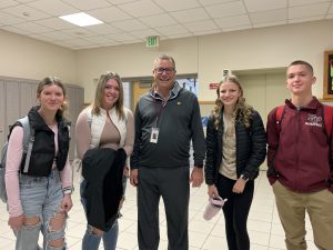 Two high school students stand on each side of Interim Jr/Sr High School Principal, Mark Fish, who is featured at the center of this photo. The group stand in a well-lit hallway of the high school and all are looking at and smiling for the camera.
