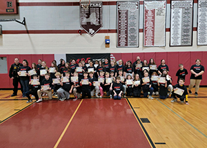 A large group of students wearing black t-shirts with the acronym "DARE" in red on their fronts stand together in a school gymnasium. All are holding certificates and looking at and smiling for the camera. 