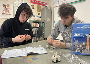 Two high school students sit side by side as a table in a school classroom. They are looking downward and are focused on their work at building small space vehicles with LEGO-type building blocks. 