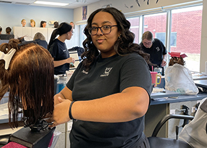 HIgh School student Nevaeh Rivers, who has long wavy dark brown hair and is wearing a black short sleeved shirt and eyeglasses, looks at and smiles slightly for the camera. Nevaeh is standing at a cutting station in a cosmetology classroom and is cutting the hair of a manequin. 