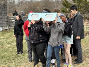 A group of teen students, holding an looking skyward through an oversized light blue pair of solar eclipse glasses, stand together outside in a field. They are all wearing sweatshirts and other cold weather clothing. There is a wooden barn and bare trees in the the background.