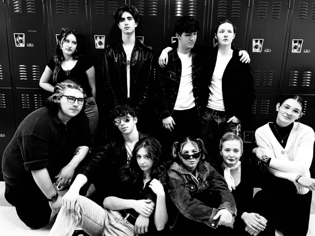 A stylized black and white photo of a group of high school students standing close together in a school hallway in front of a row of lockers. All are looking in various directions and striking a dramatic pose for the camera.