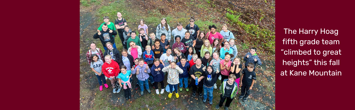 Harry Hoag fifth graders and teachers look up and smile for the camera in this aerial photo. The are all wearing hiking clothing and stand on green grass with rocky patches. The image features the phrase "The Harry Hoag fifth grade team climbed to great heights at Kane Mountain."