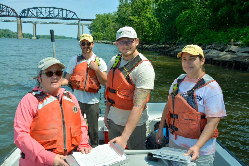 Four adults who are wearing ball caps and orange life preservers stand in a small boat on the Hudson River. All are looking at and smiling for the camera. A tree-lined shore and bridge over the water appear in the background.