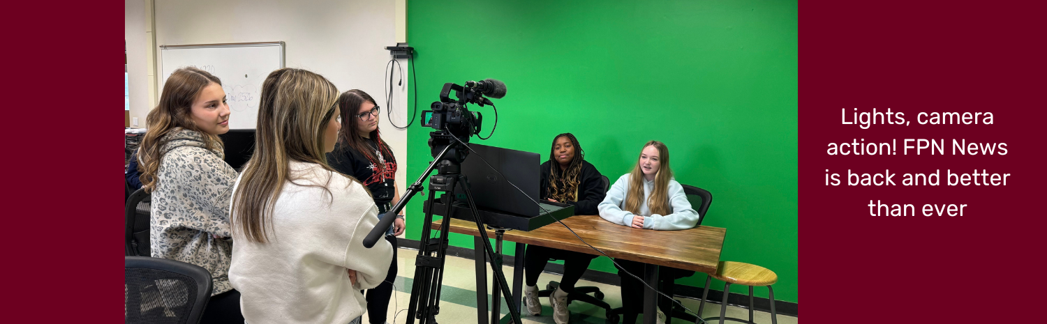 A group of high schoolers at left look through a video camera as they film a segment of FPN News. The two news anchors are seat at a table beyond the camera and in front a bright green wall.