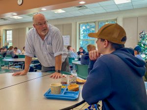 Fort Plain High School's Student Support Liaison, Will Ryan, pauses to chat with a student at a table in the school lunchroom. Students are eating and talking in groups in the background.