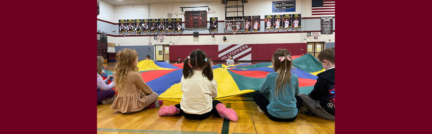 A group of young children play with a parachute while seated on the floor of the Harry Hoag elementary school gymnasium.