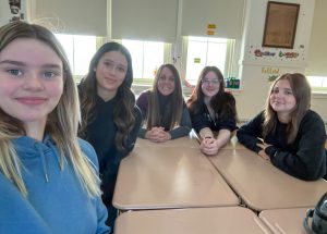 A group of teenage students sit at classroom desks with their teacher, Kolbe Gray, who is sitting at the center. They are all looking at and smiling for the camera.