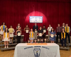 A group of high school age students wearing formal outfits and some wearing yellow drapes stand together on risers on the school stage and smile for the camera during the recent Fort Plain CSD National Honor Society induction.