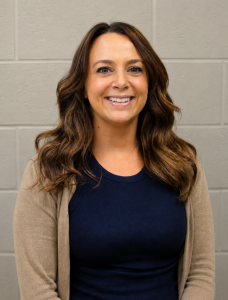 Fort Plain Technology Integration Specialist Rachel Heroth, with has long brown hair and is wearing a black top and light brown sweater, looks at and smiles for the camera.