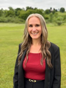A photo of Amy Bartholomew, who has long hair, is wearing a black jacket and red t shirt and smiling for the camera. Amy is standing in a green field outside with trees in the background.