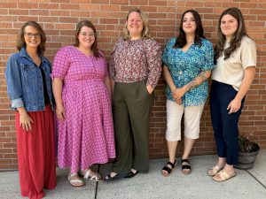 A photo of five of Fort Plain's newest faculty members seated on a district school bus. All are standing along a brick wall at Harry Hoag Elementary School and looking at and smiling warmly for the camera.