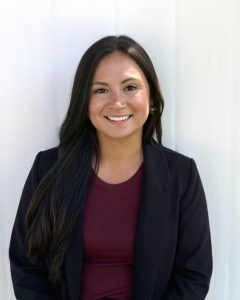 A photo of Skyler Douglas, who has long hair, is wearing a black jacket and dark red t shirt and smiling for the camera. Skyler is standing in front of a white wood paneled wall.