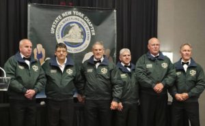 A group of former coaches, including Fort Plain's Elaine Krum, wear dark green coaches jackets and stand in a row for a photo at the Upstate New York Chapter of the Wrestling Hall of Fame induction.